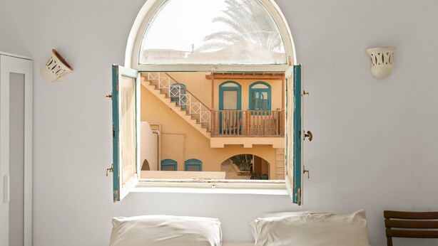 Quarto com uma grande janela em arco com vista para uma escadaria e um edifício com portas azuis.
