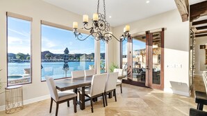 A dining area with a large table and chairs, large windows with a view of a lake and mountains, and a chandelier. - Splash Landing |  Riverfront Estate with Infinity Pool, Guest House & Private Docks (Parker)