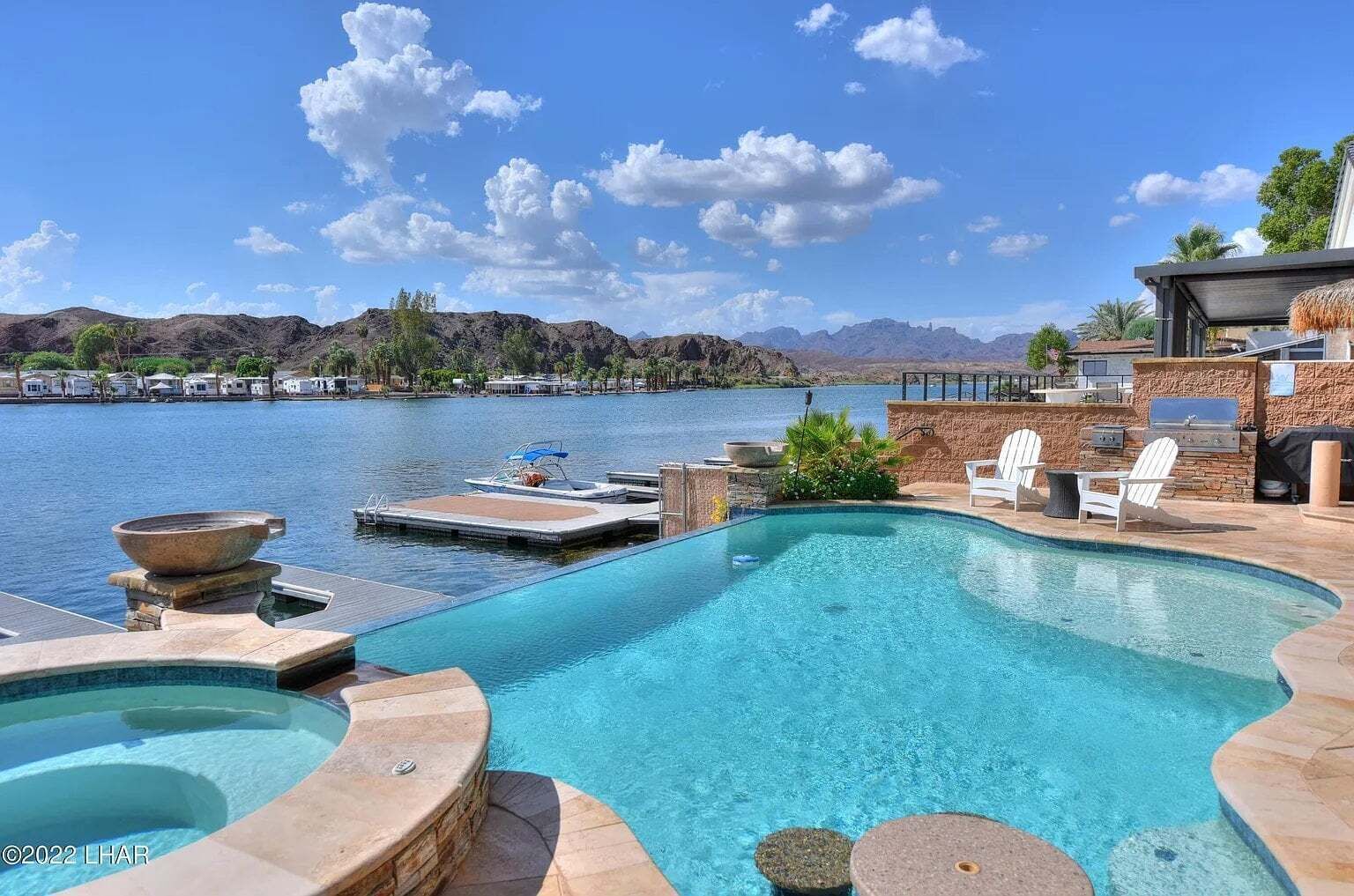 A swimming pool with a view of mountains and a lake, surrounded by lounge chairs and a barbecue area.