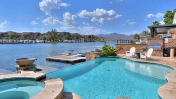 A swimming pool with a view of mountains and a lake, surrounded by lounge chairs and a barbecue area.