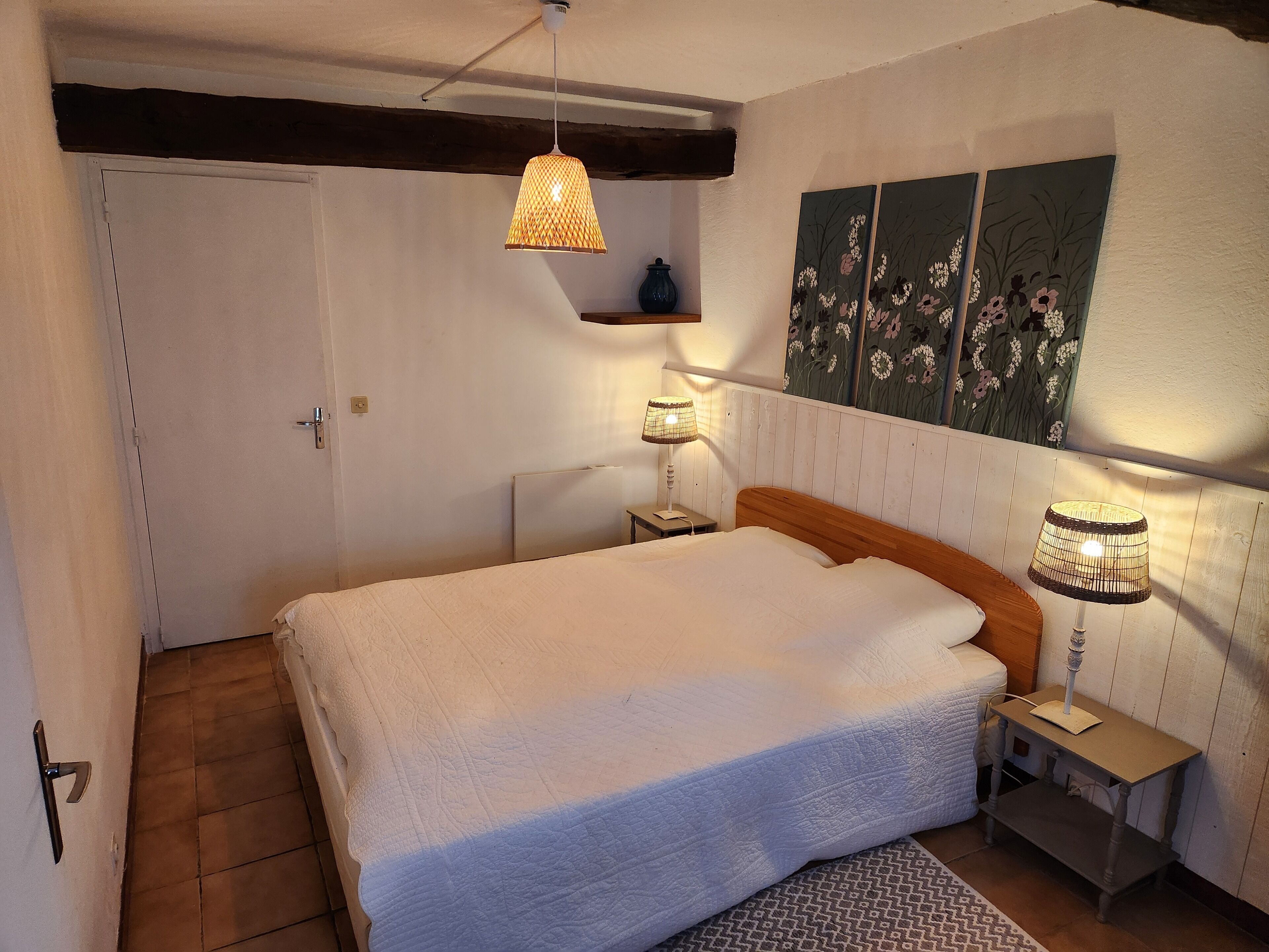 Un dormitorio con una cama grande, dos lámparas de noche, cabecera de madera, una mesita de noche pequeña y un estante fijado a la pared.