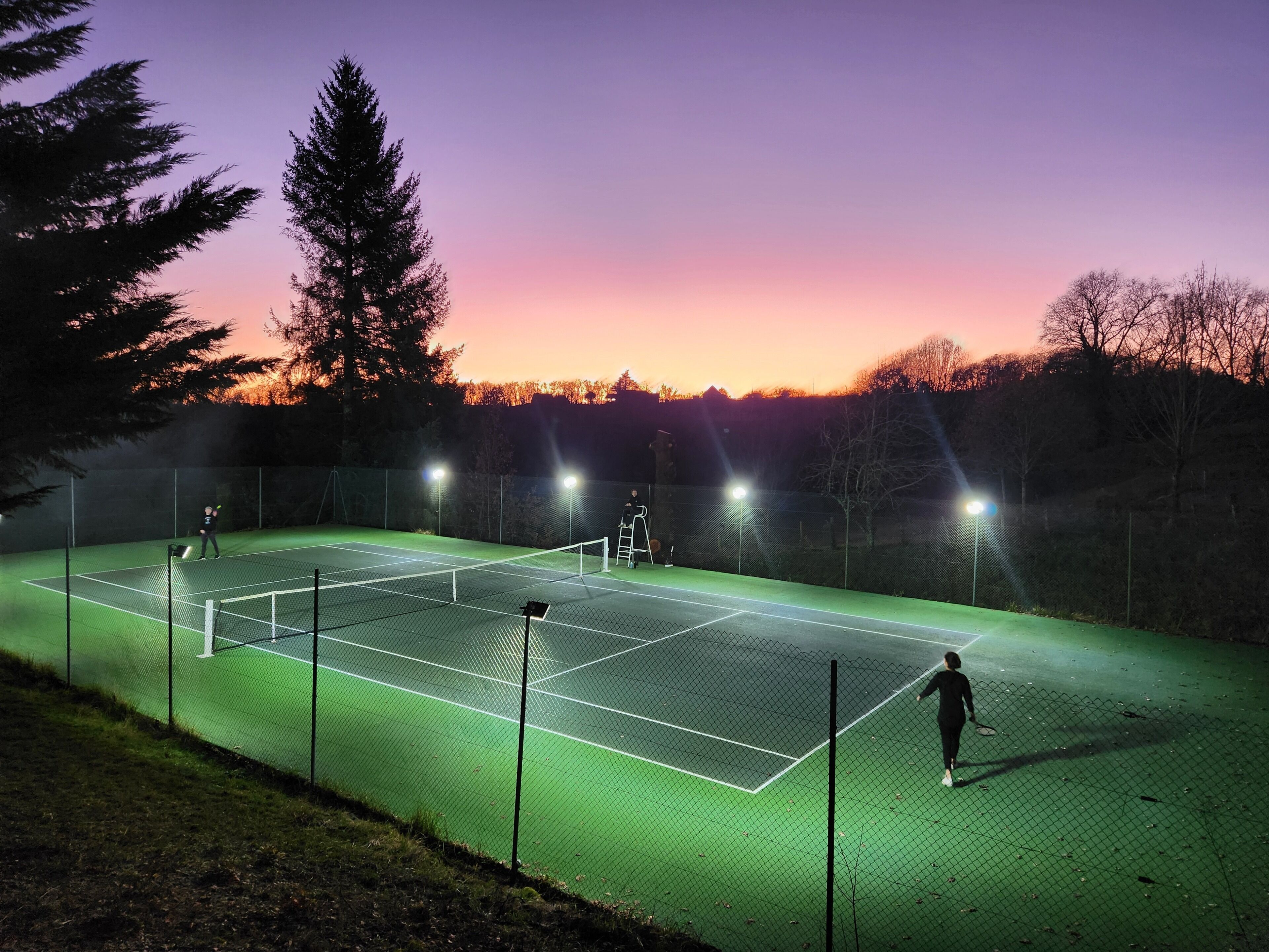 An outdoor tennis court at dusk with artificial lighting.