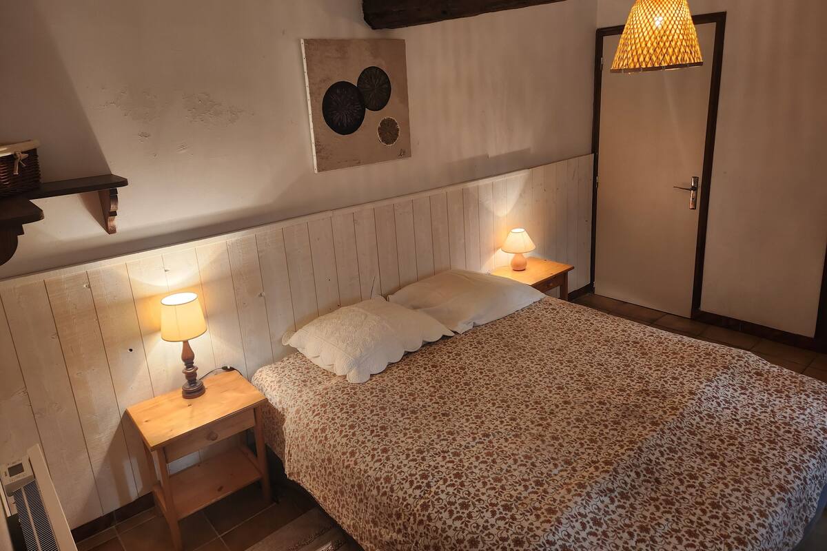 Un dormitorio con cabecera de madera, dos lámparas de noche, cama con colcha estampada, un reloj en la pared, una puerta y una lámpara colgante.