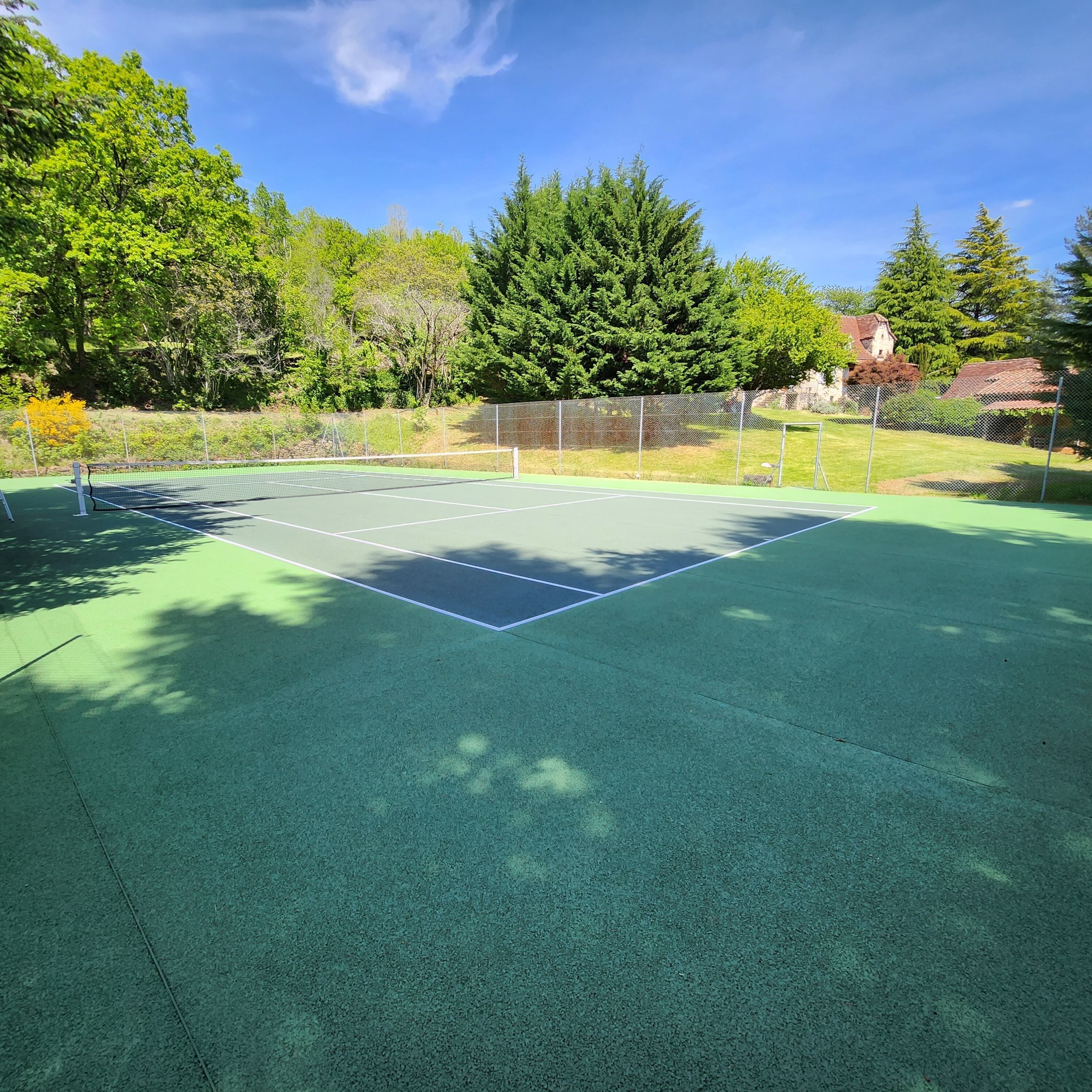 An outdoor tennis court with a green surface, surrounded by trees and a fence.