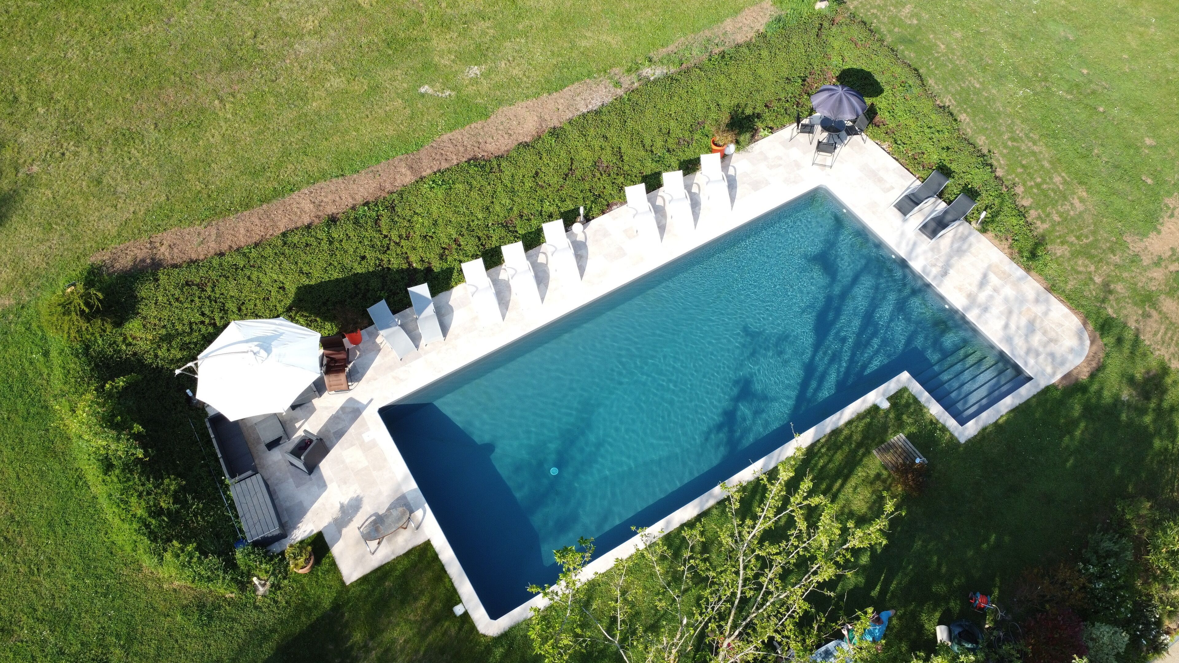 Vue aérienne d’une piscine rectangulaire entourée d’une terrasse, elle-même bordée de pelouse et d’arbres.