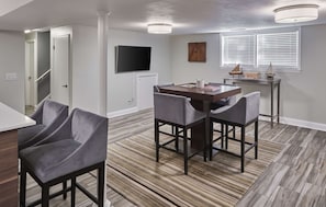 A dining area with a table and four chairs, a television mounted on the wall, and a window with blinds. - Funtana Home for 8 (Fontana)