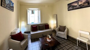 Ein Wohnzimmer mit einer Couch, zwei Sesseln, einem Couchtisch und einem Fenster mit geöffnetem Rollo.