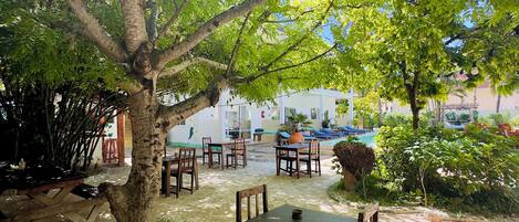 An outdoor seating area with wooden tables and chairs under a large tree, surrounded by greenery and a pool in the background.