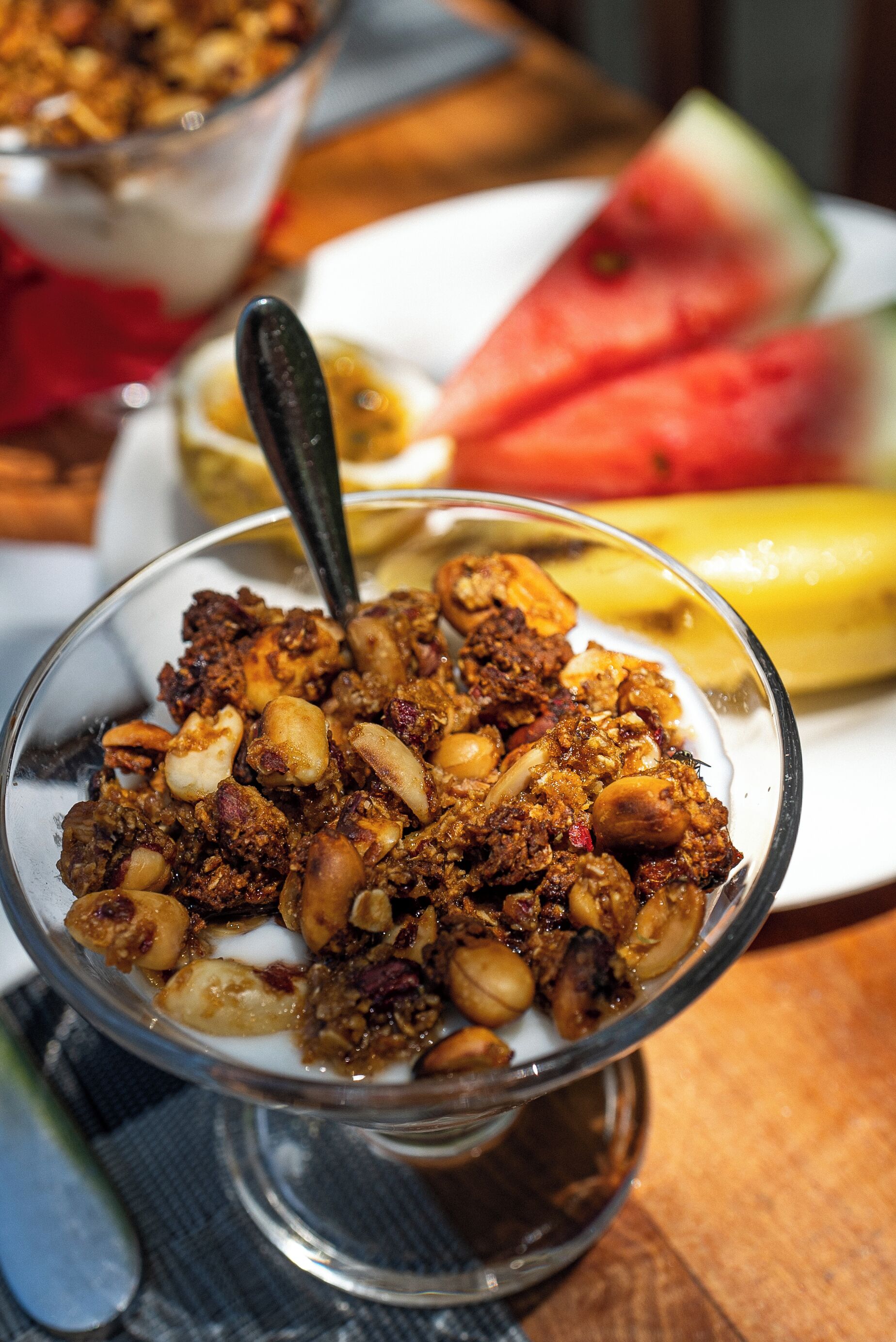 A dessert bowl with granola and nuts, a spoon, and a plate with watermelon and banana in the background.