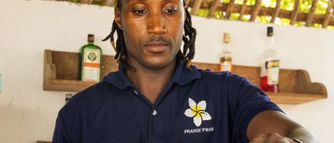 Un barman prépare des boissons dans un bar doté d’un comptoir en bois et d’un décor tropical.
