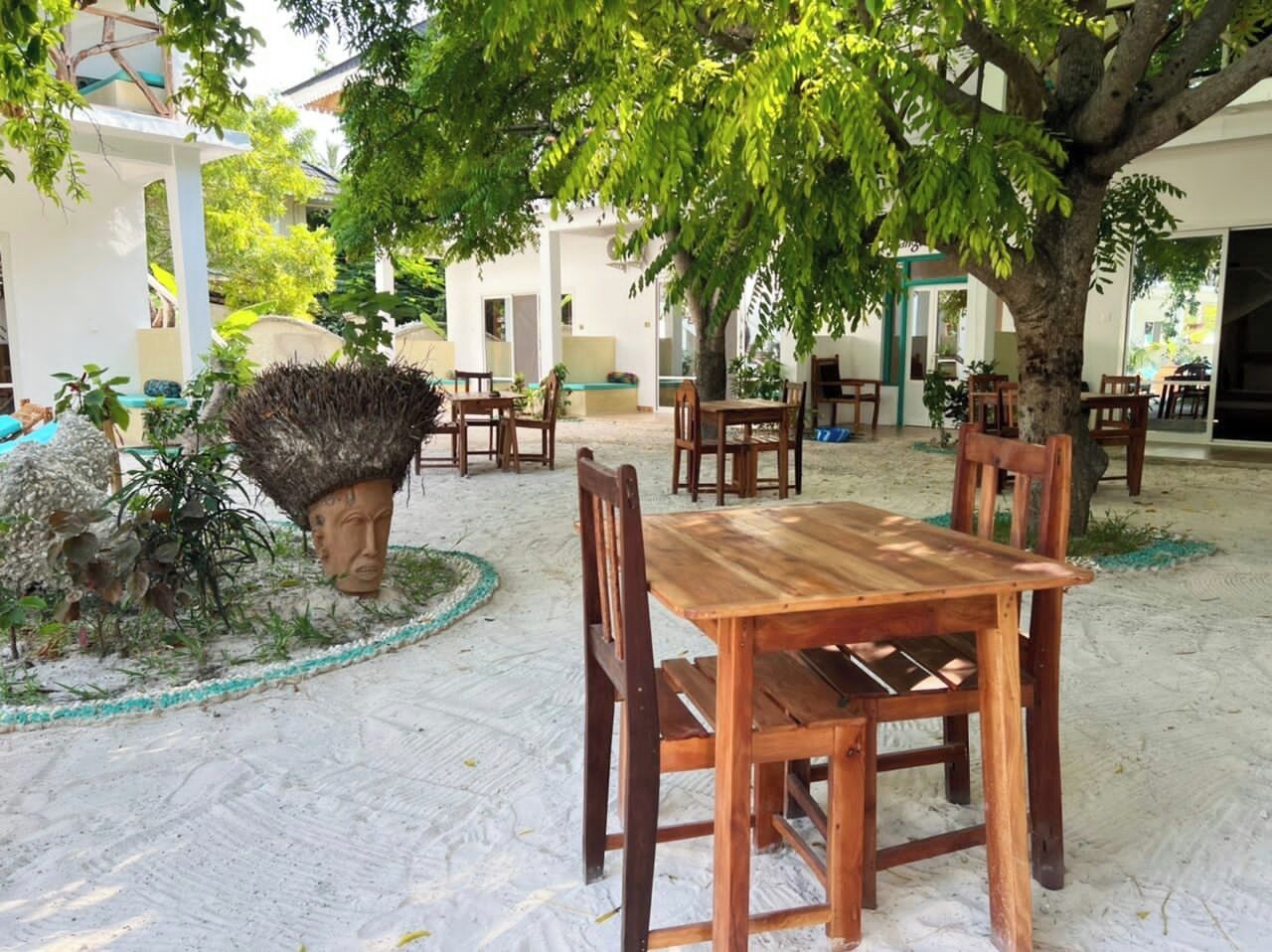 Un área de comedor al aire libre con mesas y sillas de madera, rodeada de árboles y suelo arenoso.