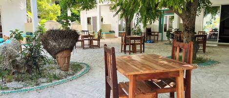 Un área de comedor al aire libre con mesas y sillas de madera, rodeada de árboles y suelo arenoso.