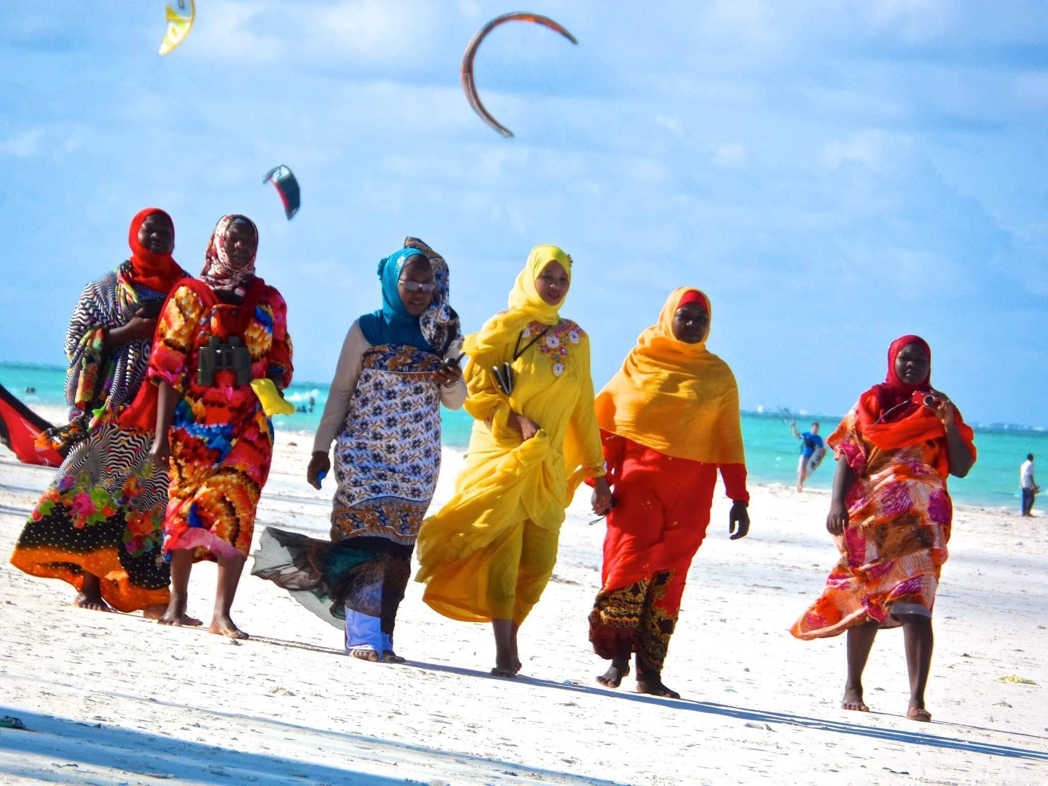 A group of people walking on a beach with kites in the sky.