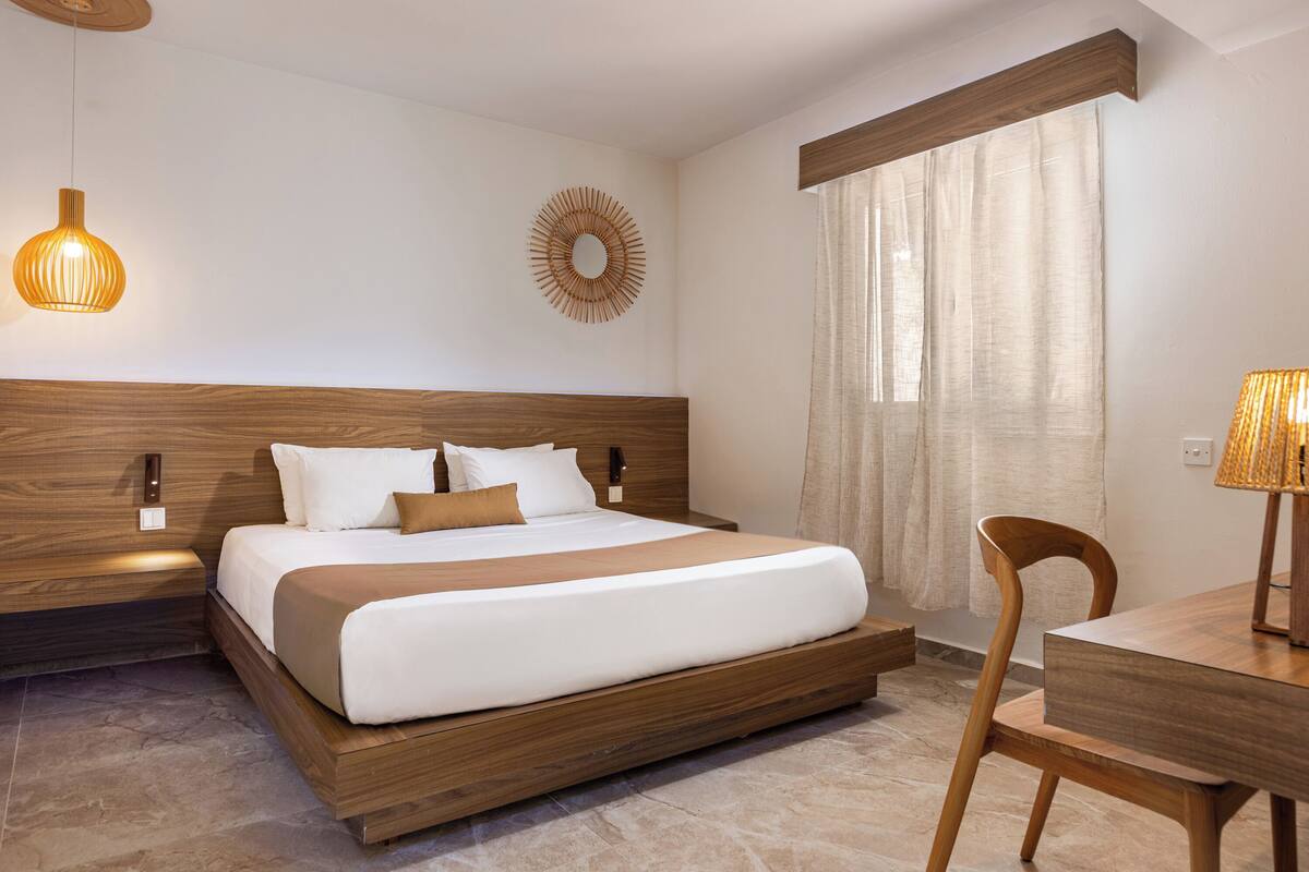 Una camera d'albergo moderna con una testiera in legno, un letto grande, un comodino con una lampada e vista all'esterno attraverso una finestra con tende.
