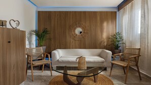 A modern living room with a glass coffee table, wicker chairs, a white sofa, and a wooden accent wall with a circular mirror. - So Apartments (Ayia Napa)