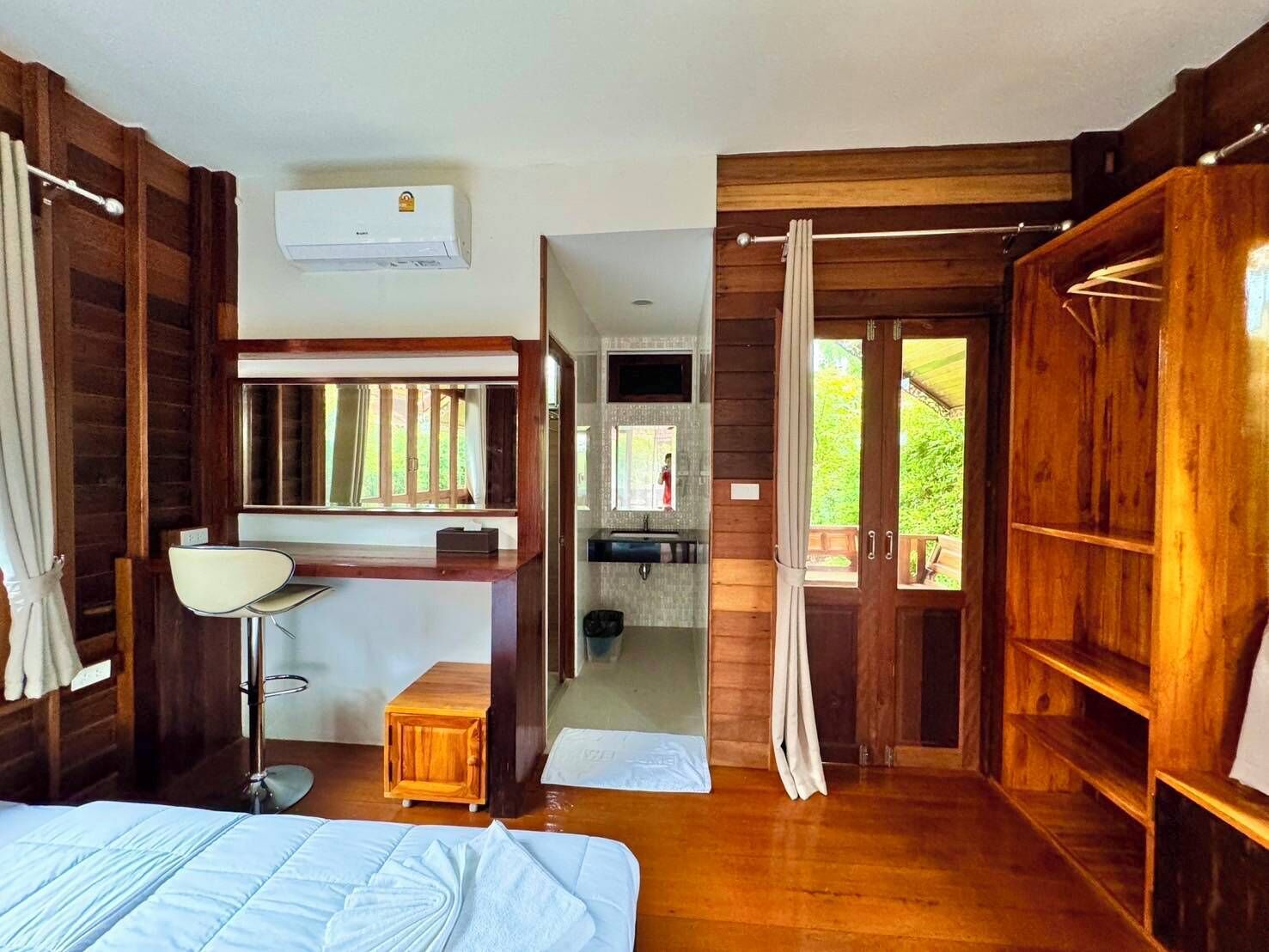 A wooden room with a bed, a desk, a mirror, a window, and a shower area.
