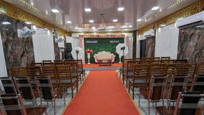 A conference room with a red carpet, rows of wooden chairs, a stage with a backdrop, and air conditioning units. - Selvam Residency (Thoothukudi)