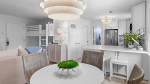 A modern kitchen with white cabinetry, a central island, and a dining area with a round table and wicker chairs. - Happily Ever After on 30A w/FREE golf cart New Condo (Santa Rosa Beach)