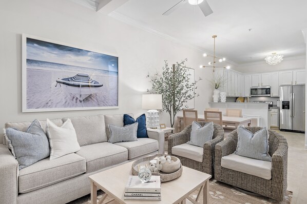 A modern living room with a large beach-themed artwork, a sofa set, and a dining area with wicker furniture. - NEW rental in Seagrove on 30A w/Golf Cart! (Santa Rosa Beach)
