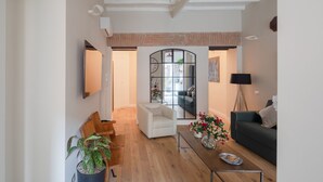 A modern living room with a wooden floor, a white sofa, a coffee table, and a dining table. There is a brick wall and a large window. - THE BREAK - PIAZZA DI SPAGNA MAISON DELUXE TERRACE (Roma)
