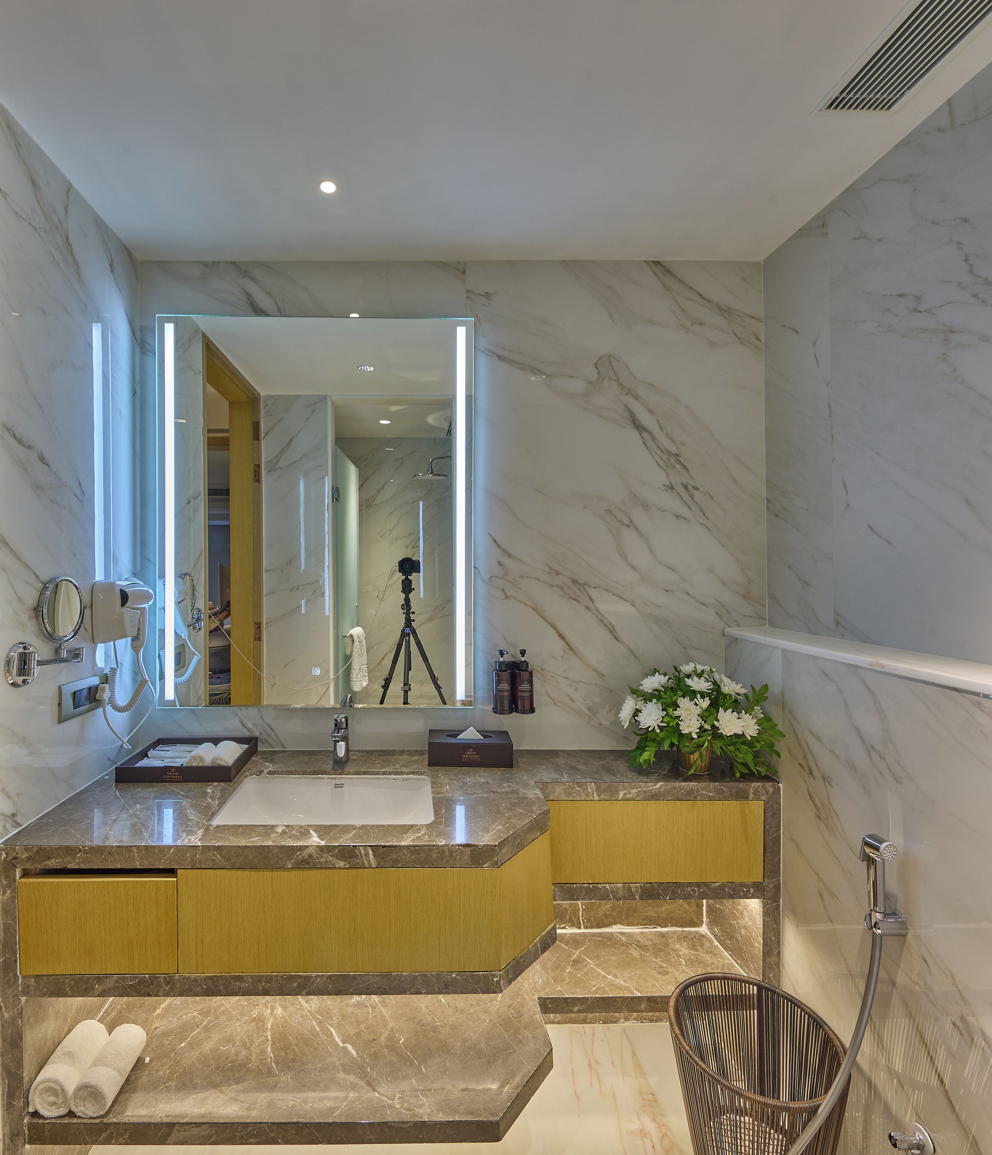A modern bathroom with marble countertops, a large mirror, a camera on a tripod, and a shower area.