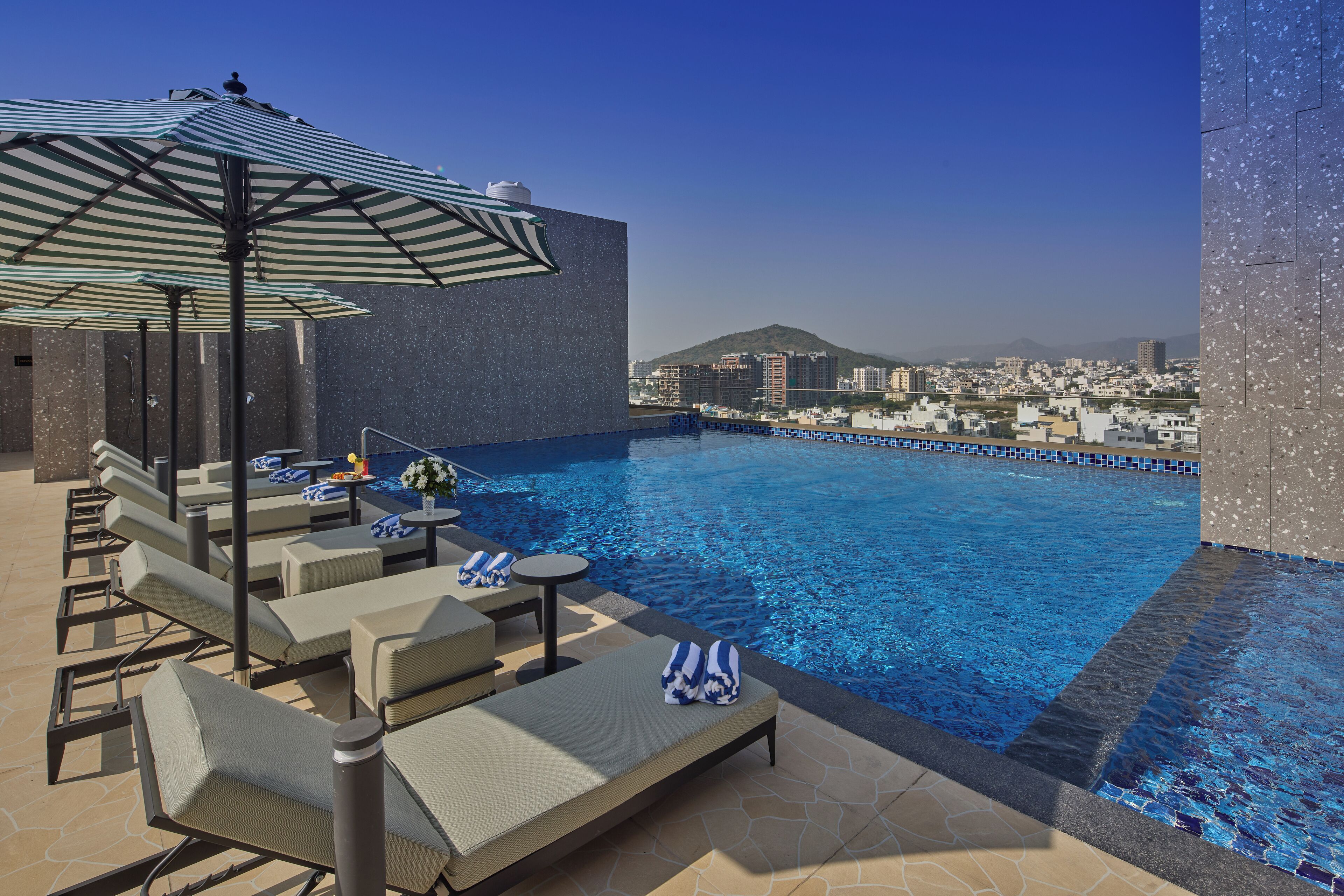 Un área de piscina en la azotea con sillones de descanso, una sombrilla a rayas y vistas de la ciudad.