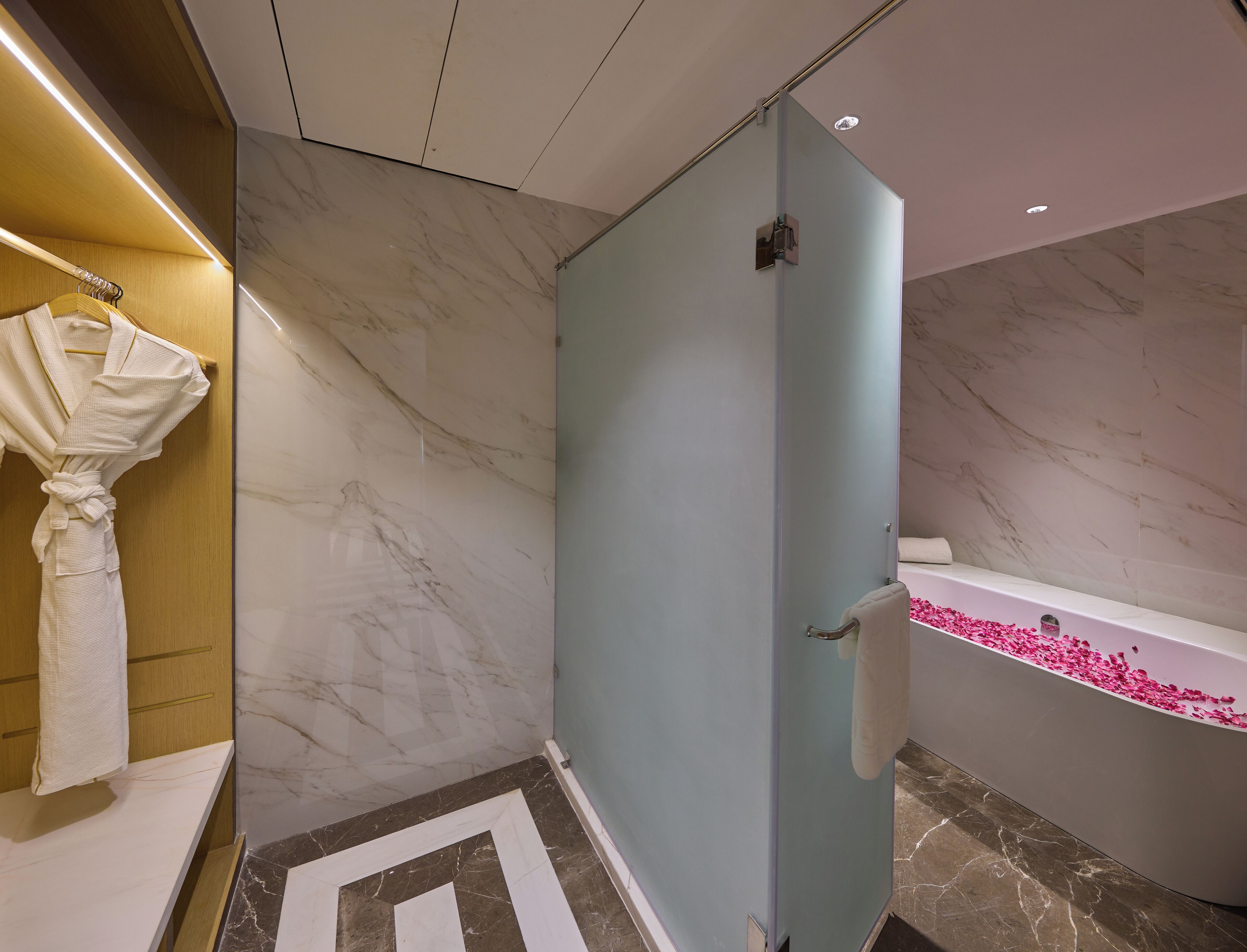 Une salle de bain moderne avec un sol en marbre, une douche en verre, une baignoire remplie de pétales de rose et un peignoir suspendu à un porte-serviettes en bois.