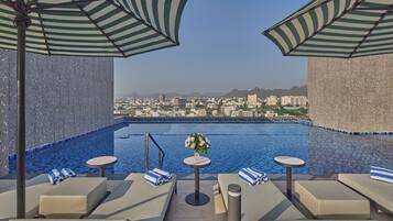 A rooftop pool area with lounge chairs, striped umbrellas, and a view of the cityscape.