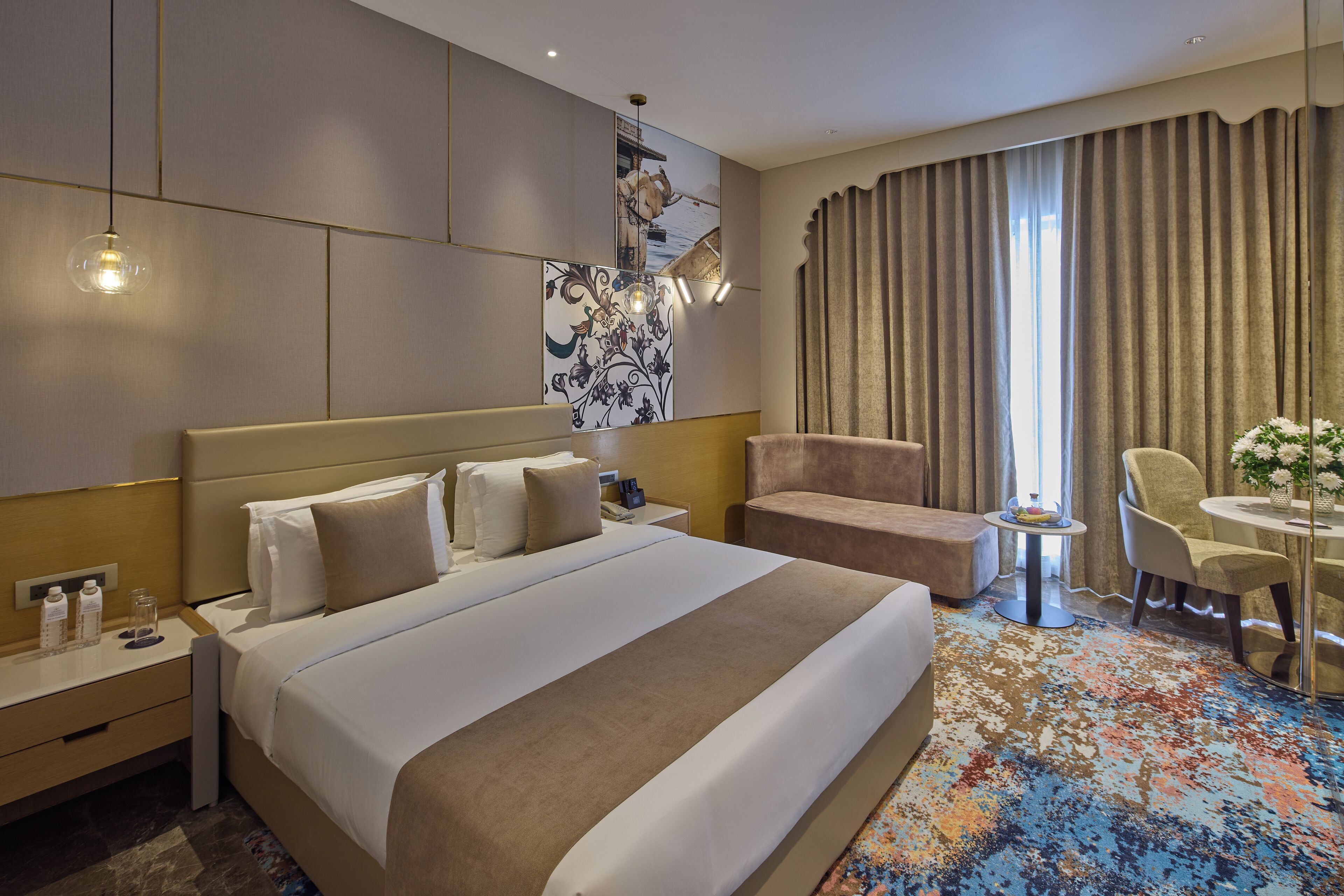 A hotel room with a large bed, a desk with a lamp, a chair, and a small table with a vase.