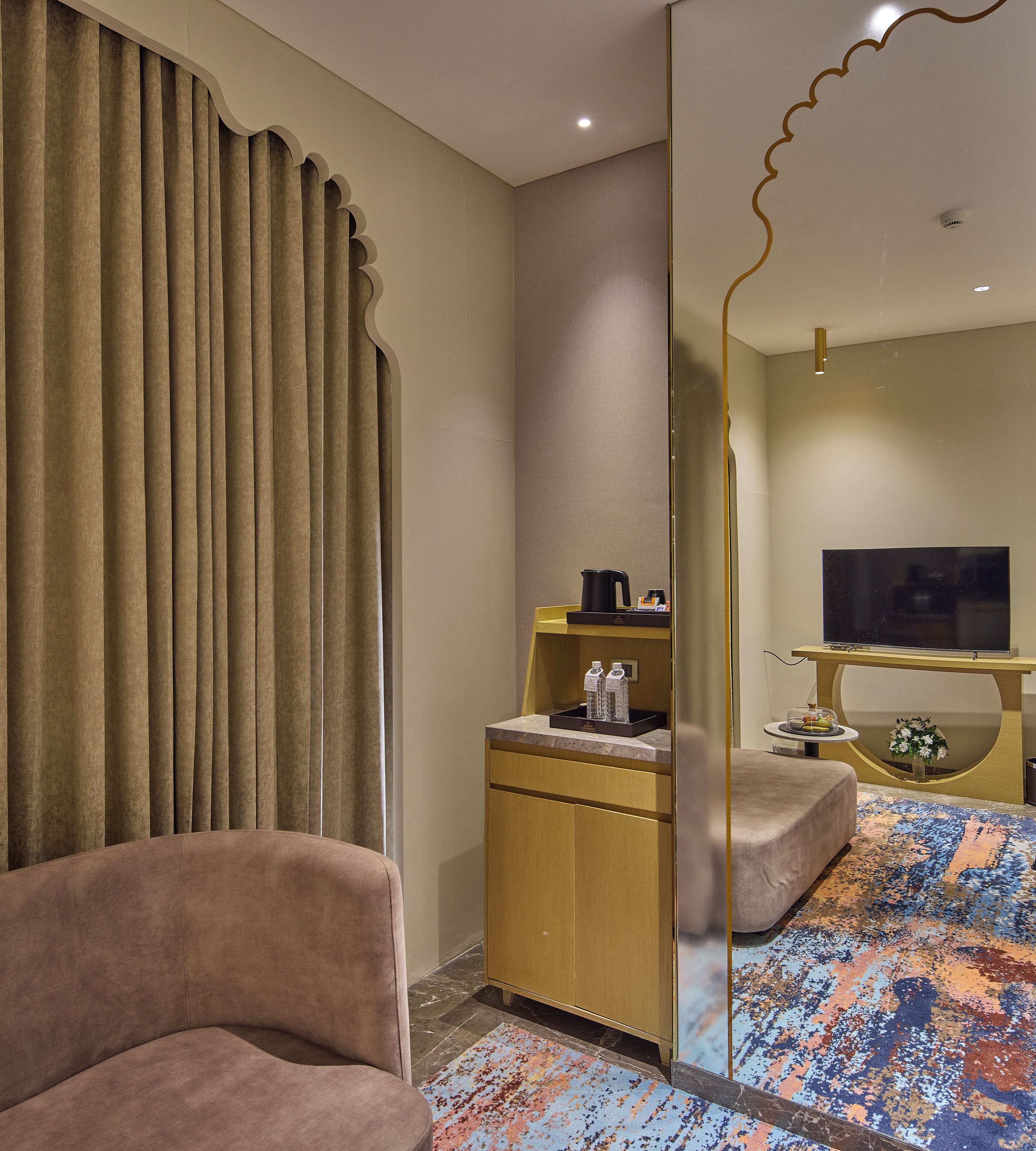 A hotel room with a sofa, a desk with a TV, a minibar, and a patterned rug.