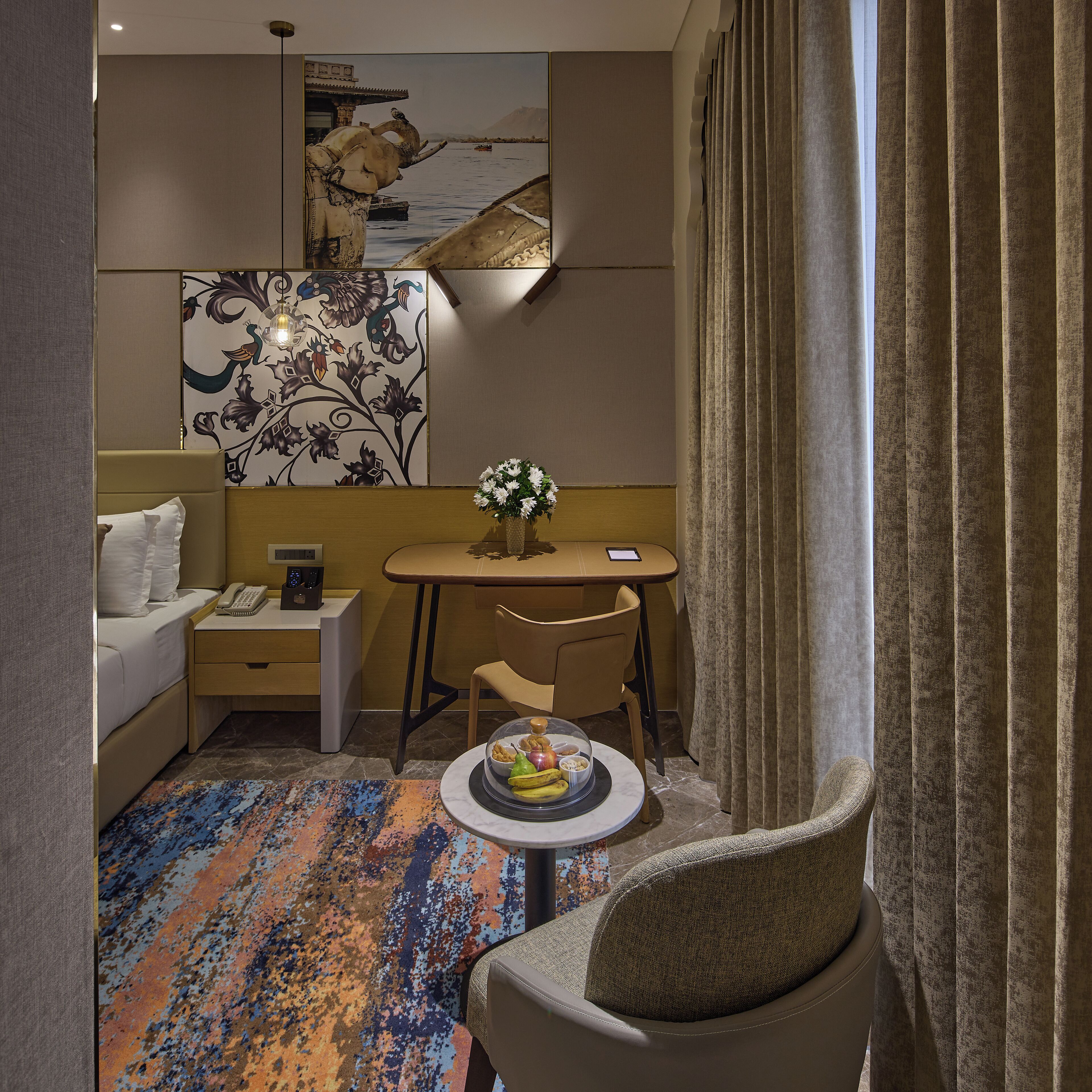 A hotel room with a bed, a desk, a chair, a small table with a fruit bowl, and a colorful rug.