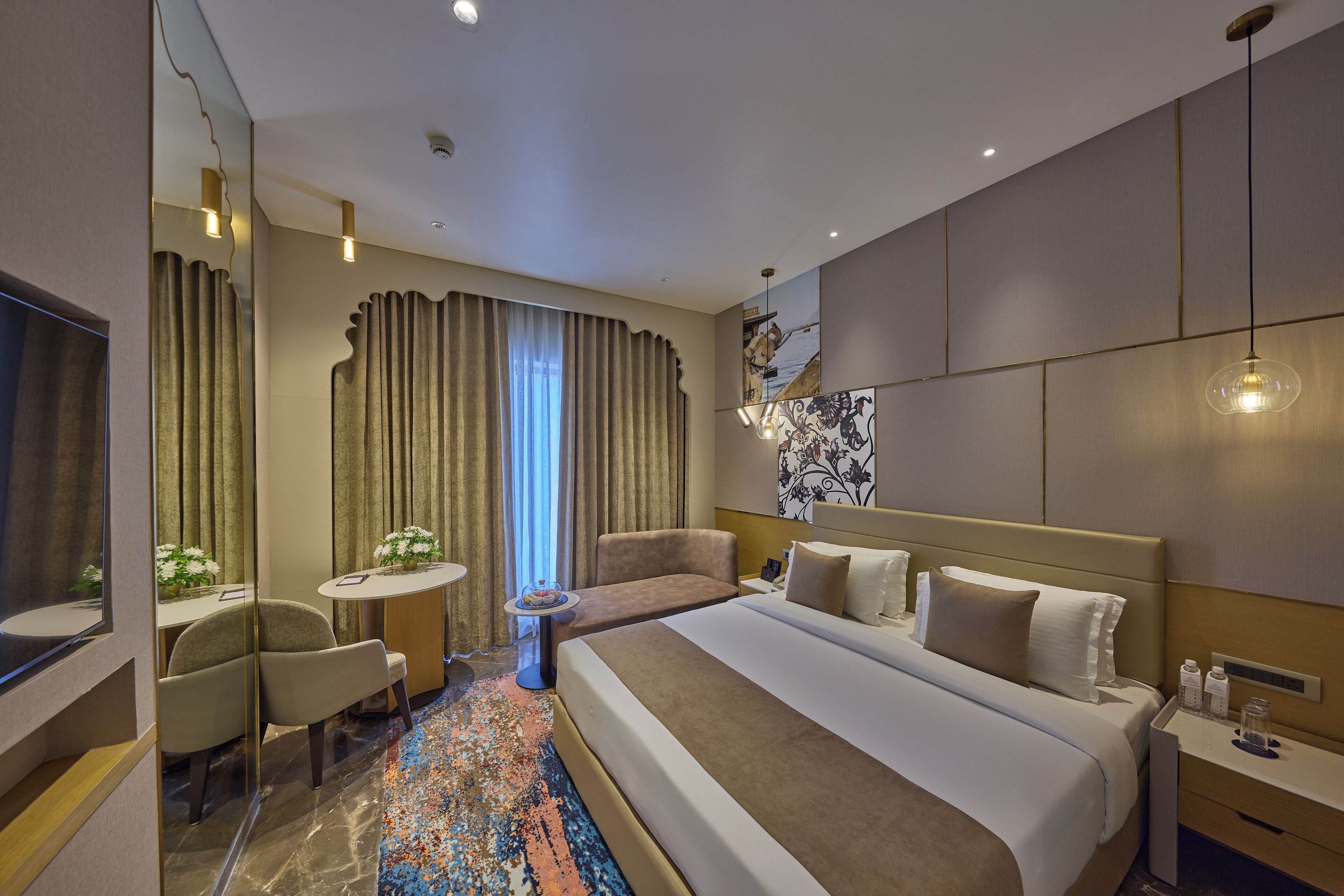 A modern hotel room with a large bed, a sitting area with a chair and table, a small round table with a vase of flowers, and a large window with curtains.