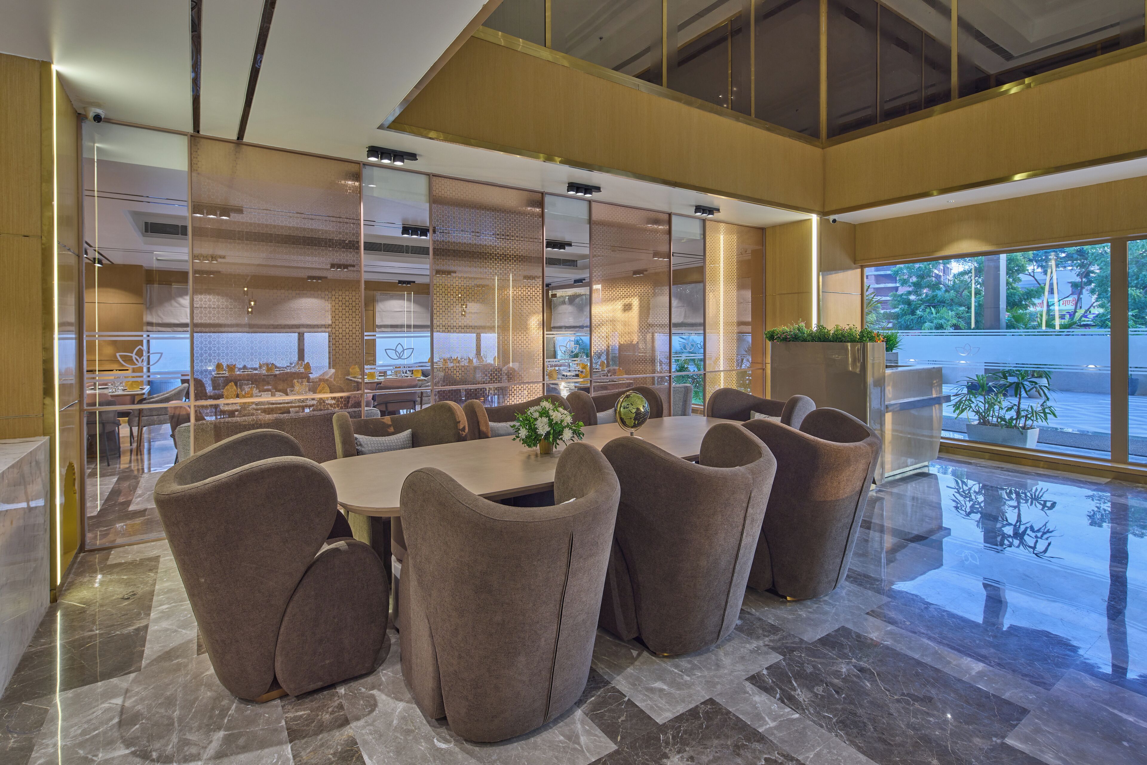 A modern dining area with a large table and six upholstered chairs, a glass partition, and a view of the outdoors.