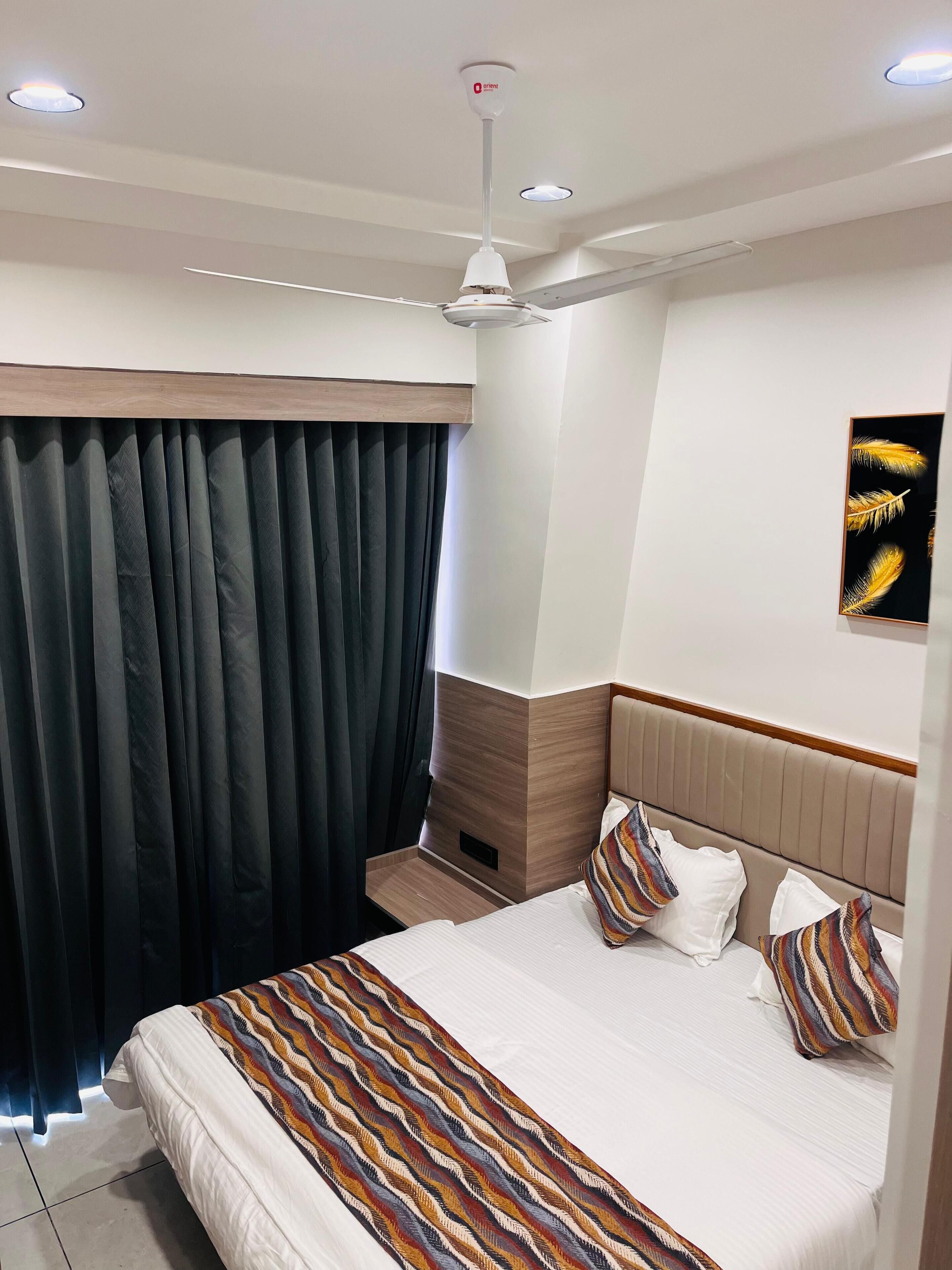 Une chambre d’hôtel avec un lit, un ventilateur de plafond, une fenêtre avec des rideaux et une œuvre d’art accrochée au mur.