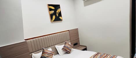 Une chambre d’hôtel bien rangée, avec un lit, une table de chevet, un climatiseur fixé au mur, un ventilateur de plafond et une œuvre d’art encadrée accrochée au mur.