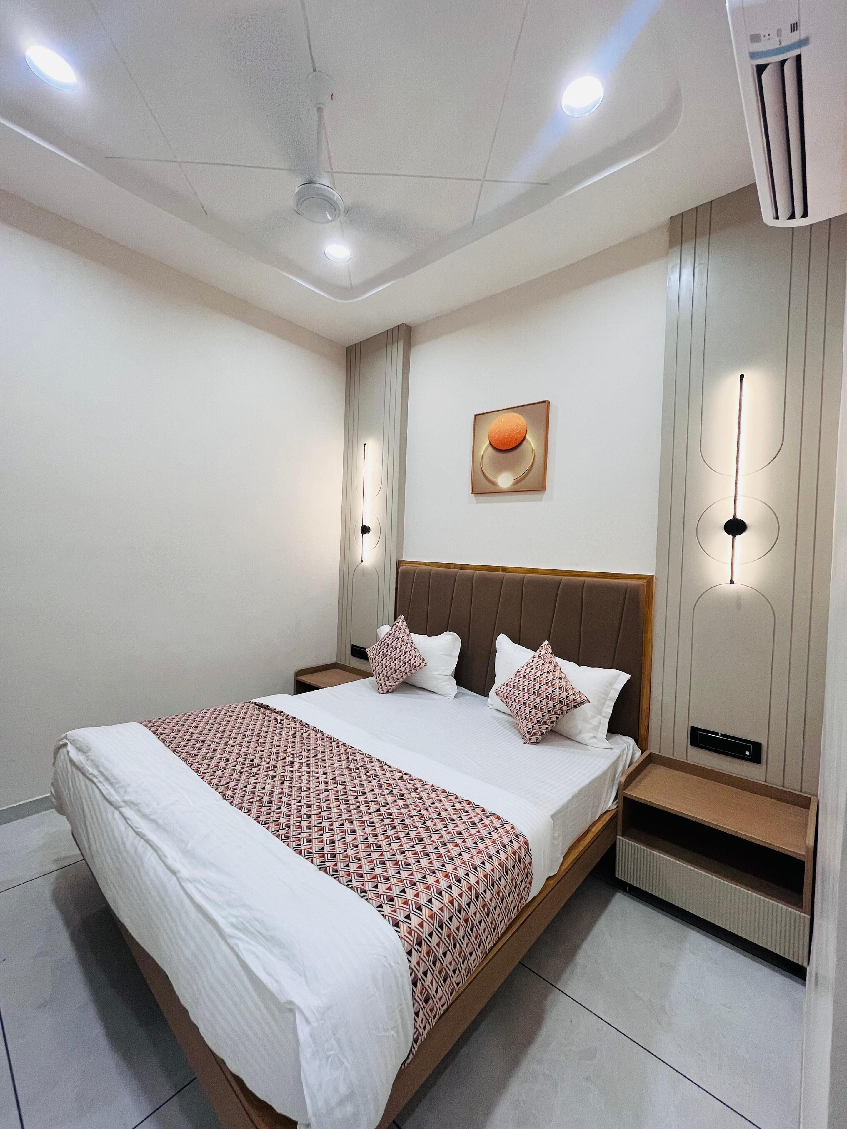 A modern hotel room with a large bed, a bedside table, a wall-mounted air conditioner, and a ceiling light fixture.