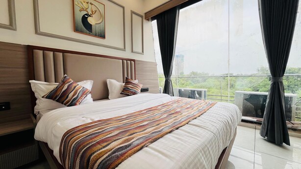 A hotel room with a large bed, a headboard, a painting on the wall, and a view of greenery through the window.