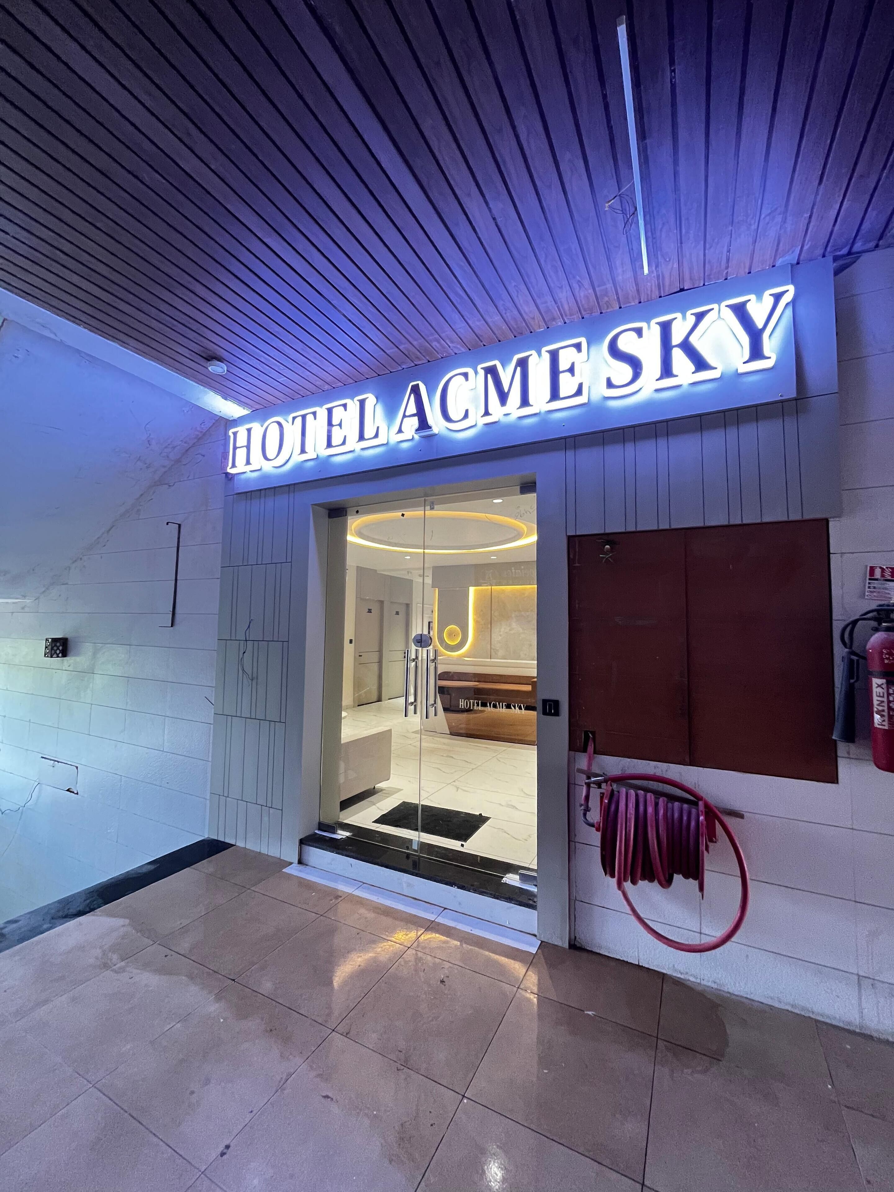 The entrance of HOTEL ACME SKY with a modern design, including a glass door and a visible reception area.