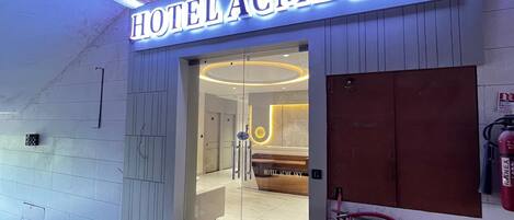The entrance of HOTEL ACME SKY with a modern design, including a glass door and a visible reception area.