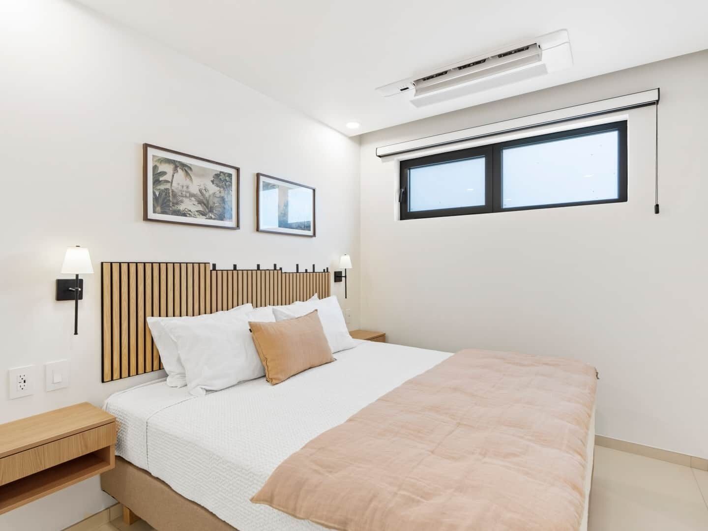 A modern bedroom with a bed, bedside tables, wall-mounted air conditioning, and two framed pictures.