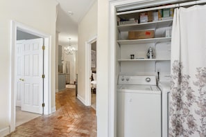A laundry room with a washing machine, shelves with boxes, and a door leading to a hallway.