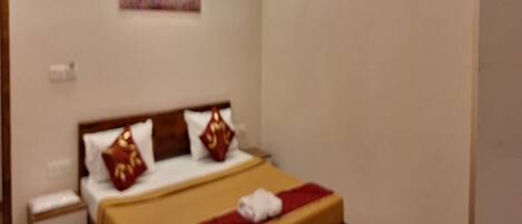 A hotel room with a bed, a nightstand, an air conditioning unit, and a painting on the wall.