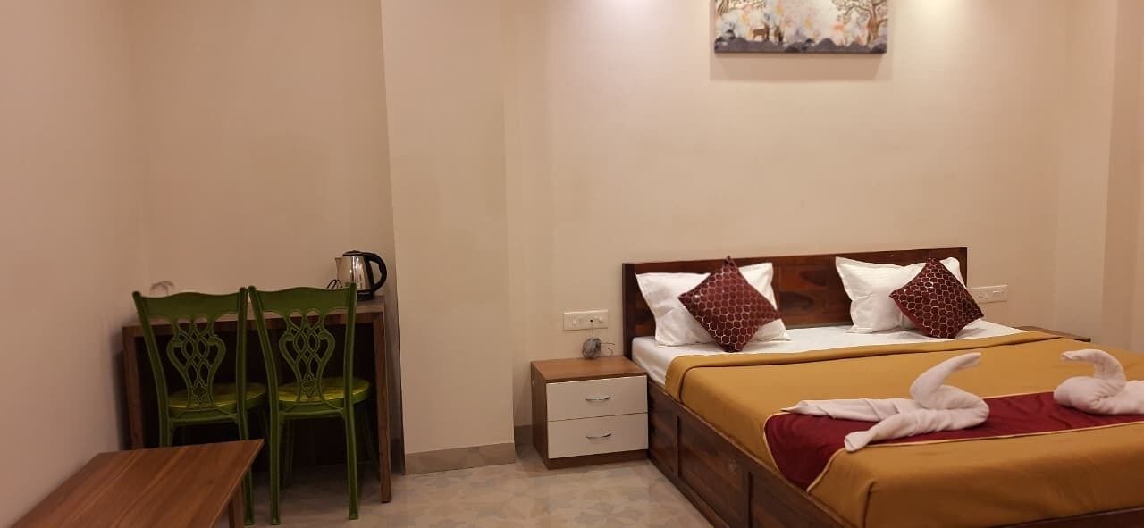 Habitación de hotel con una cama, dos sillas verdes, una mesita, una mesilla de noche y un cuadro en la pared.
