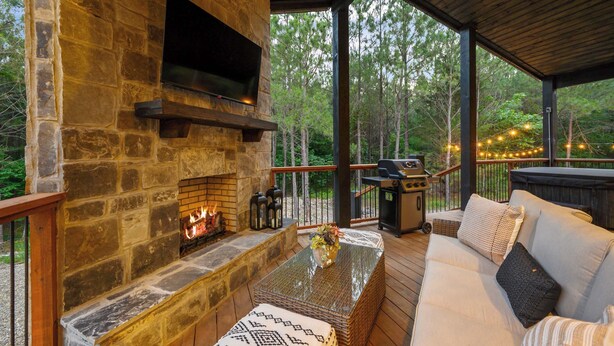 A stone fireplace with a TV, outdoor seating, and a deck with string lights. - New! At Ease Romantic Honeymoon Cabin Hot Tub (Broken Bow)