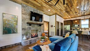 A living room with a stone fireplace, blue sofa, and a television mounted on the wall. - New! At Ease Romantic Honeymoon Cabin Hot Tub (Broken Bow)