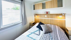 A bedroom with a bed, a window with curtains, a bedside table, a nightstand, and built-in wooden shelving above the bed. - Mobile home - 2 bedrooms - air conditioning - Enclosed terrace - Locsweethome (Les Mathes)
