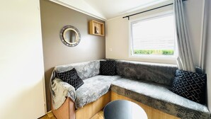 A compact living space with a sofa, a small round table, a mirror, and a window with curtains. - Mobile home - 2 bedrooms - air conditioning - Enclosed terrace - Locsweethome (Les Mathes)
