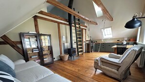 Un salon de style industriel, avec un plancher en bois, un canapé, un fauteuil, une bibliothèque, une échelle et un coin cuisine équipé d’une cuisinière.
