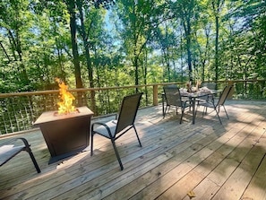 Eine Holzterrasse mit Feuerstelle, Sitzgelegenheiten im Freien und Blick auf einen Wald.