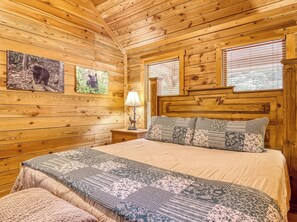 Un dormitorio de cabaña de madera con una cama, una mesita de noche, una lámpara y dos ventanas.
